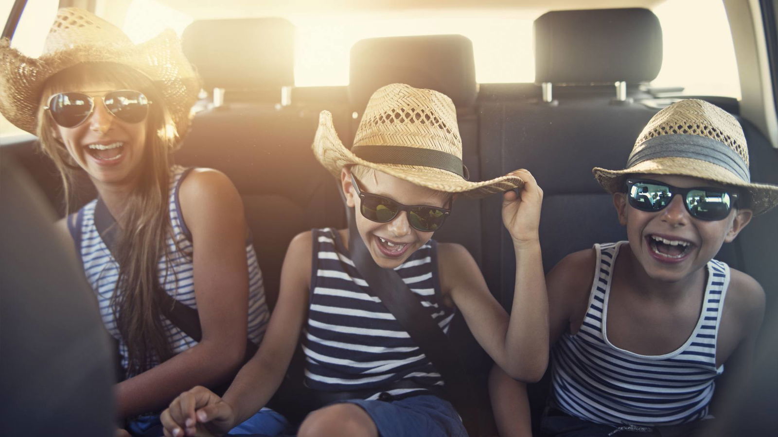 Drei Kinder mit Hüten und Sonnenbrillen sitzen auf der Rückbank eines Autos und lachen sich kaputt.
