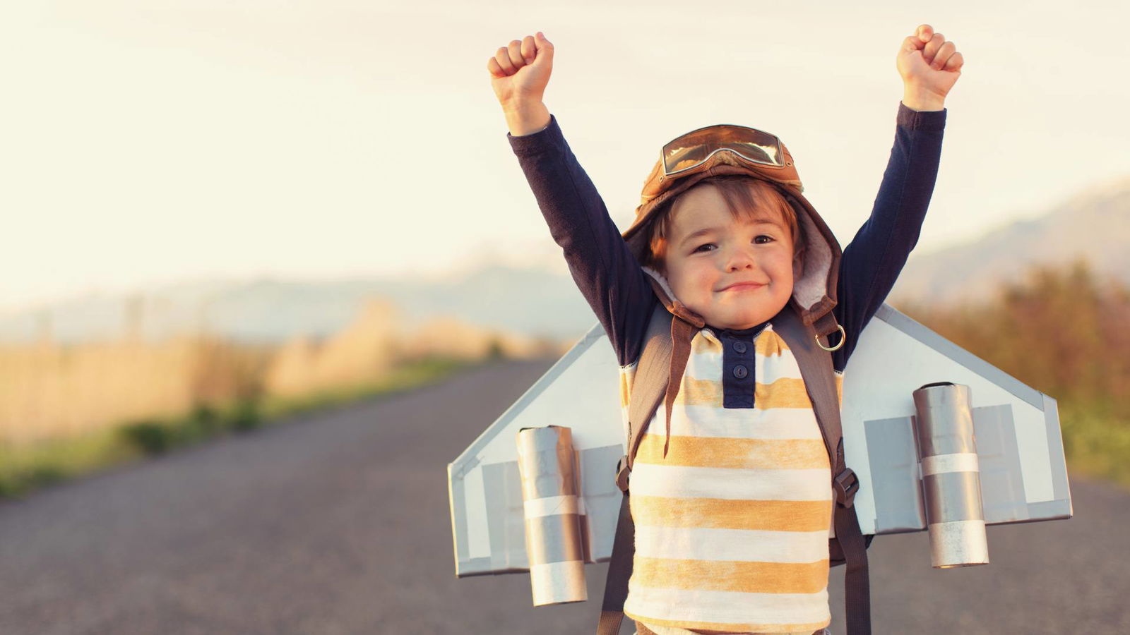 Ein kleiner Junge hat einen selbst gebastelten Flieger auf dem Rücken und reißt die Arme hoch.