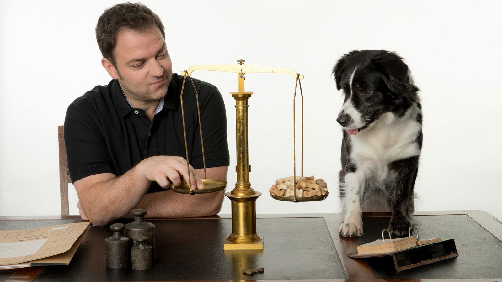 Ein Mann und ein Hund sitzen gemeinsam an einem Tisch auf dem eine Waage steht. Der Mann legt Leckerlies in die Schalen und wird dabei vom Hund beobachtet.