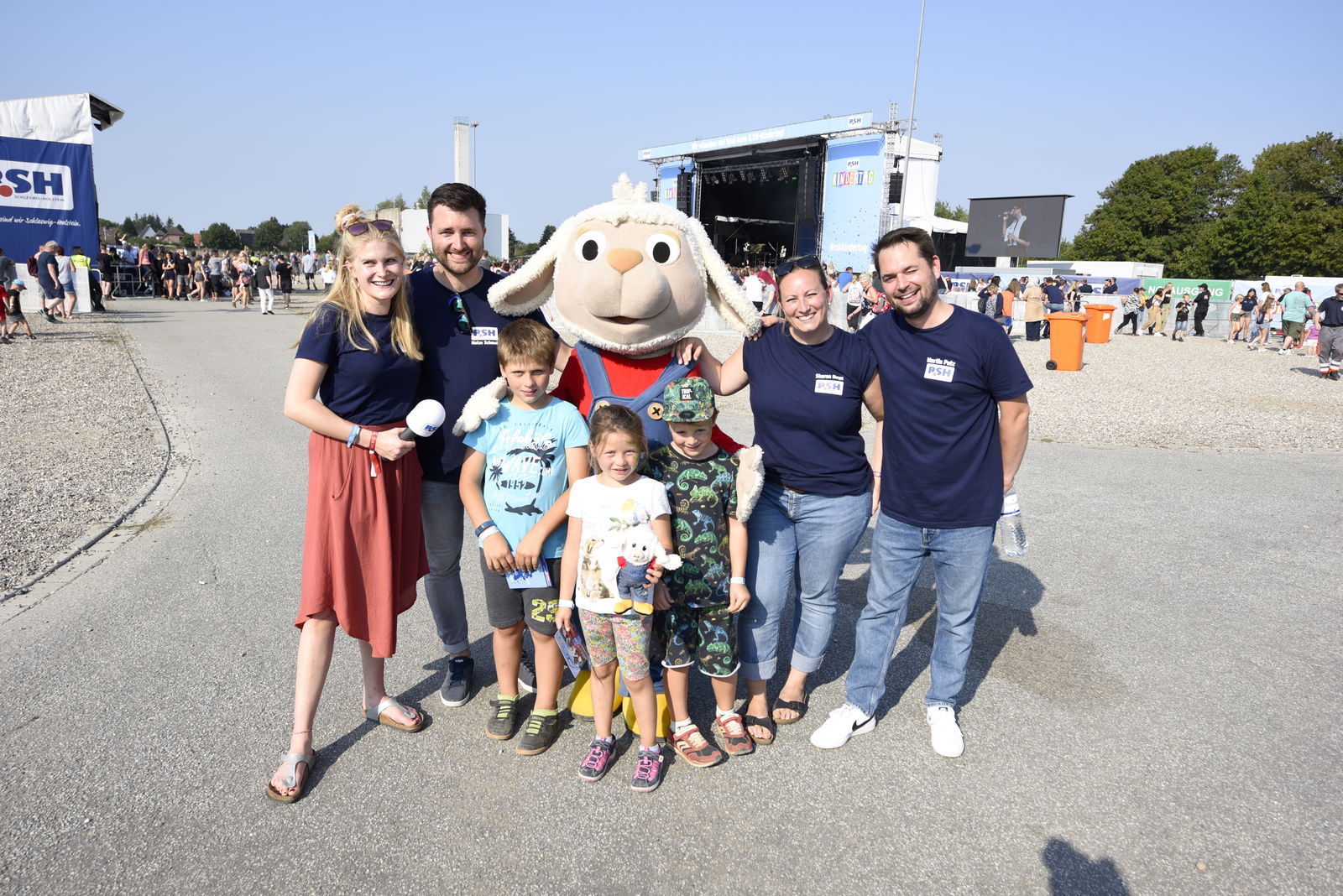 Drei kleine Kinder stehen vor einem Schafmaskottchen. Links neben dem Maskottchen stehen ein Mann und eine Frau. Rechts daneben ebenfalls. Gemeinsam lachen sie in die Kamera.   