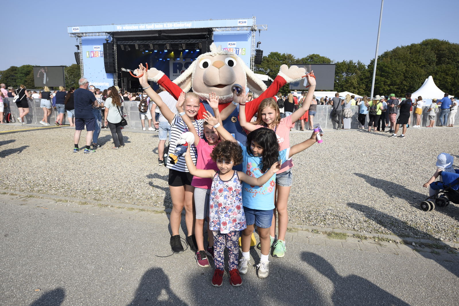 Fünf kleine Kinder stehen vor einem Schafsmaskottchen und schauen in die Kamera. Gemeinsam heben sie die Hände in die Luft.  