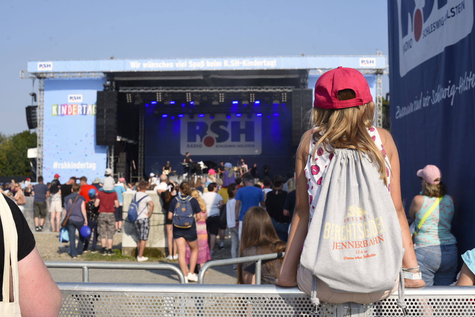Ein Kind mit langen Haaren, einem Rucksack und einer roten Kappe schaut auf eine Bühne und hat dabei der kamera den Rücken zugewandt.