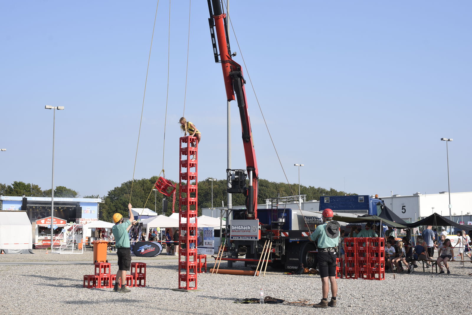 Mehrere Getränkekisten bilden einen Stapel. Auf diesen Stapel klettert eine Person die durch ein Seil, das an einem Kran hängt, gesichert ist. Vor dem Kistenstapel stehen zwei Personen. 