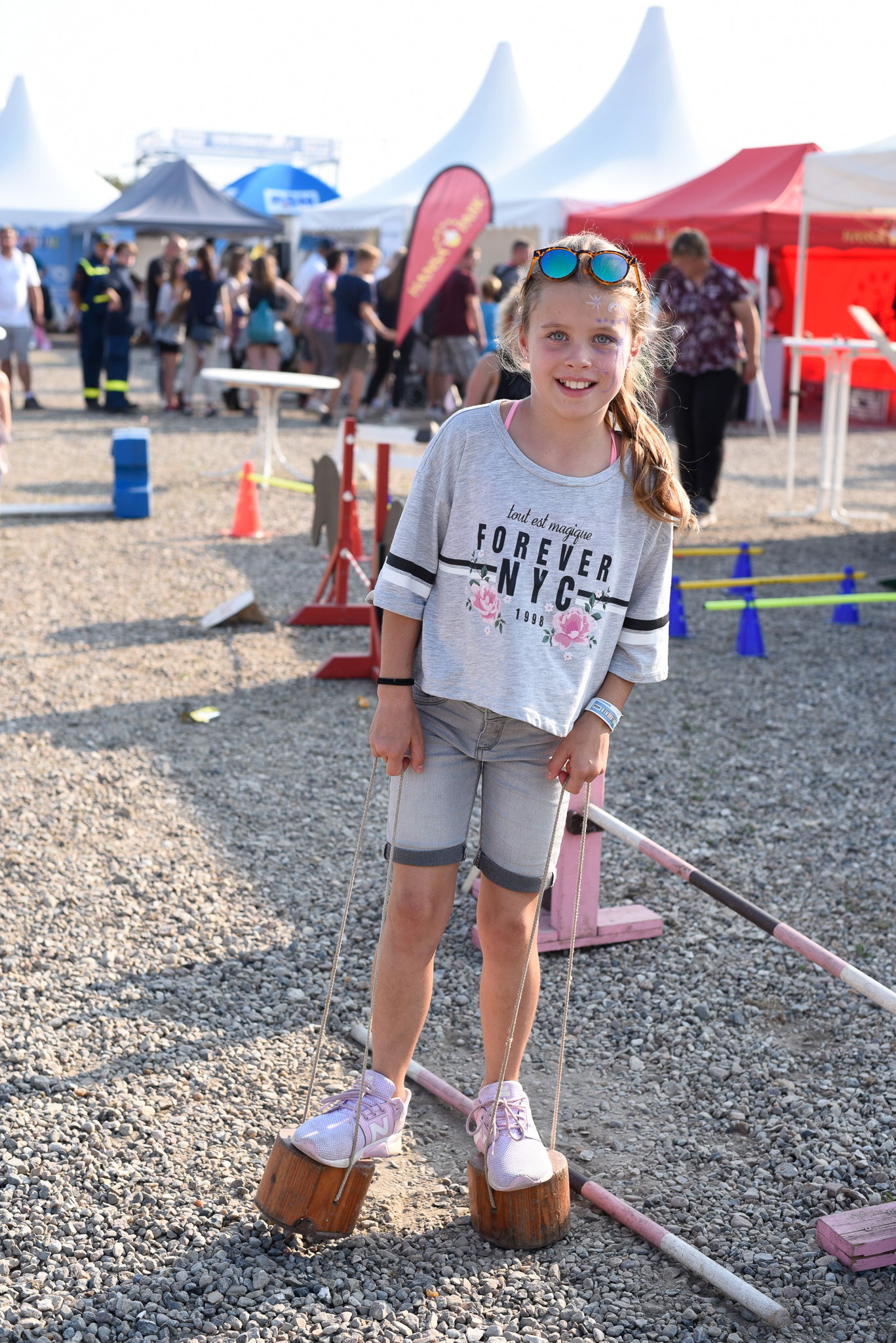 Ein kleines Mädchen balanziert auf Elefantenfüssen (Stelzen), auf einer Kiesfläche und lacht in die Kamera. 