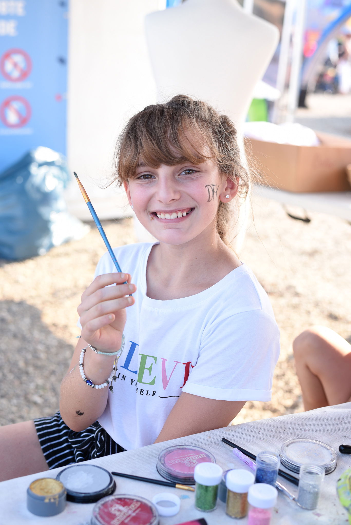 Ein Mädchen sitzt an einem Tisch mit Schminkutensilien, hält einen Pinsel in der rechten Hand und lächelt in die Kamera.