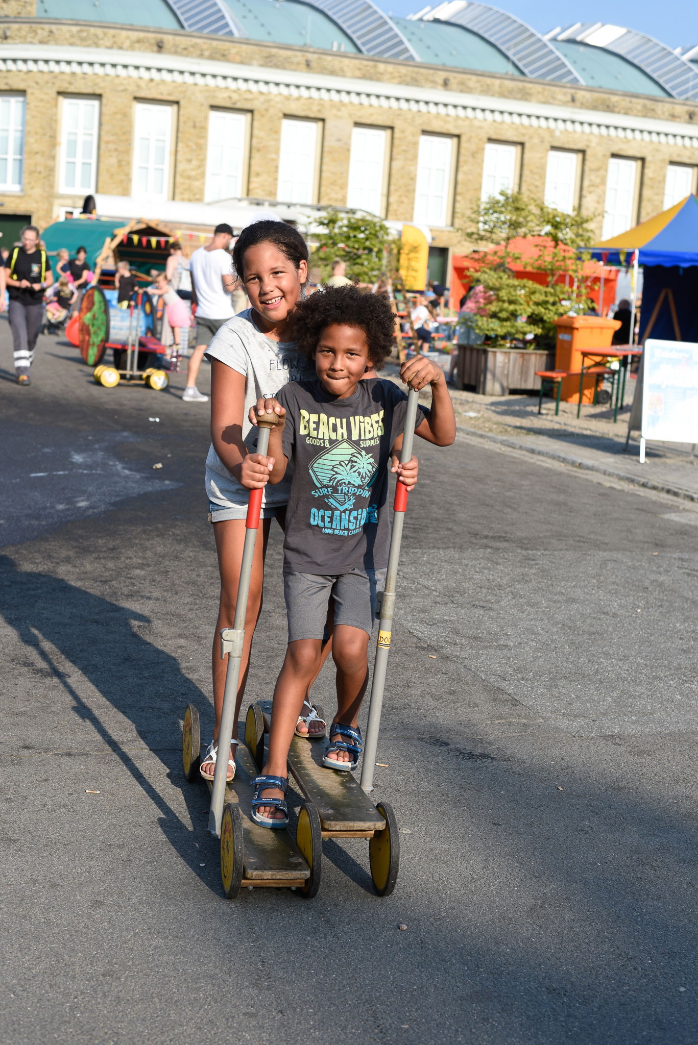 Zwei kleine Mädchen stehen gemeinsam auf einem Tretroller und versuchen damit zu fahren.