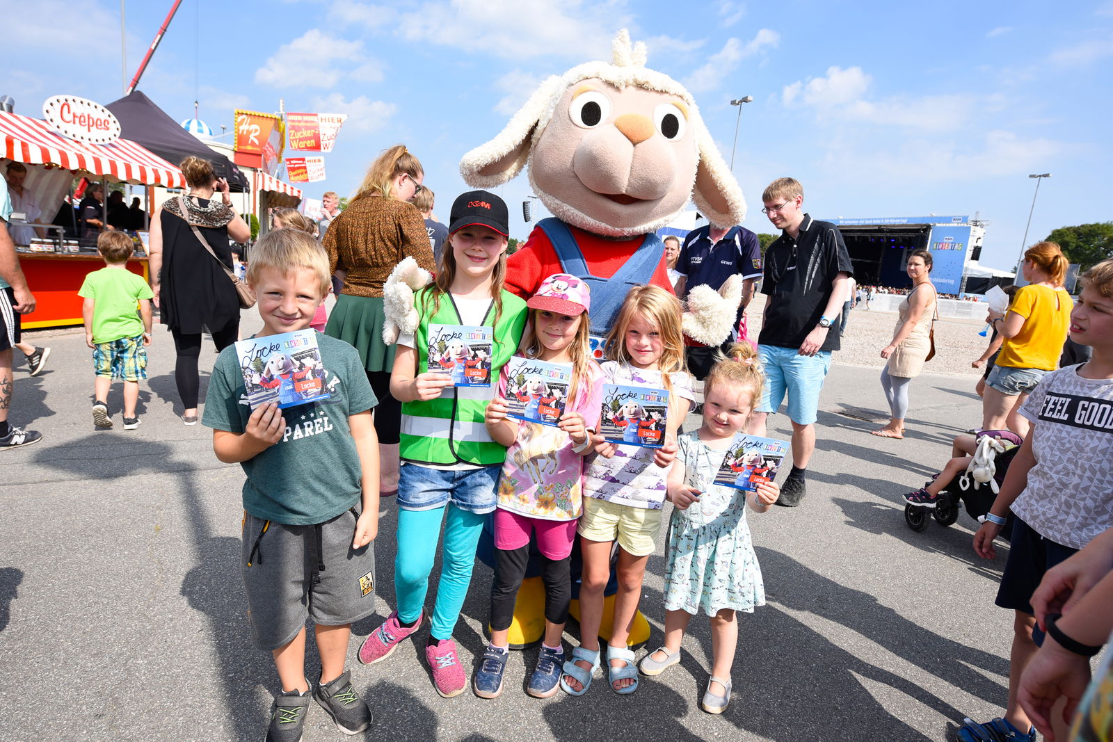 Fünf kleine Kinder stehen vor einem Maskottchen in einem Schafskostüm. Alle Kinder halten eine Autgrammkarte des Maskottchen in der Hand und lächeln in die Kamera. 