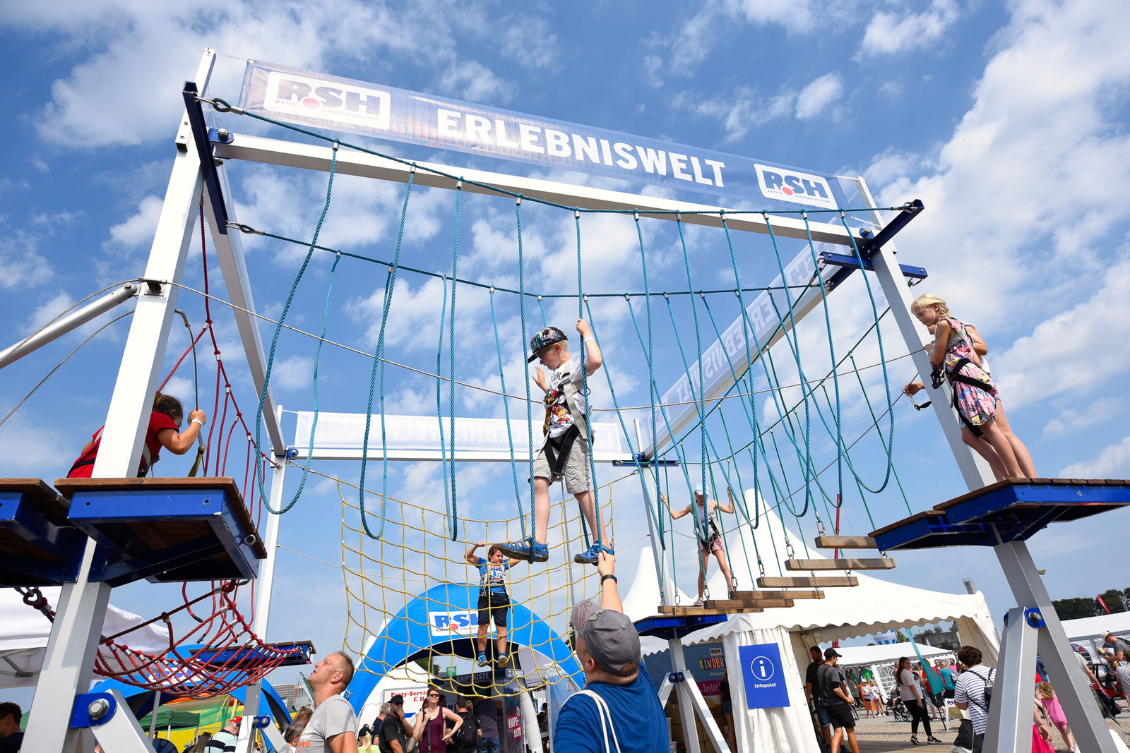 Ein Junge geht von einer blauen Plattform auf der rechten Seite, auf der eine Frau steht, über eine Hängebrücke zu einer weiteren blauen Plattform auf linken Seite. Auf ihr steht ein Mann. Das Kind ist durch Seile gesichert.