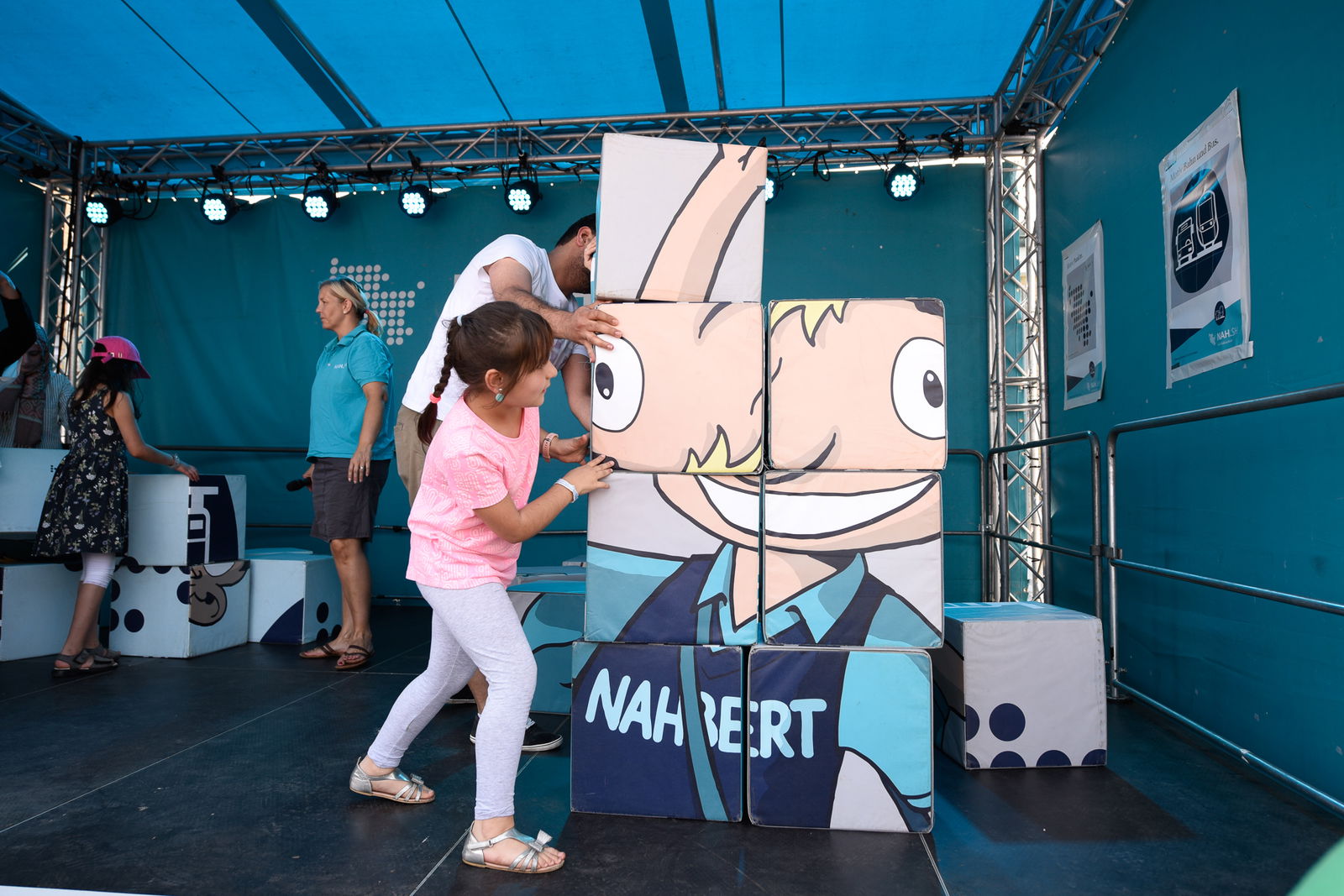 Ein junges Mädchen steht links neben einem riesen Puzzle aus Blöcken, auf dem eine Comicfigur zu sehen ist. Das Mädchen versucht die Blöcke korrekt anzuordnen.