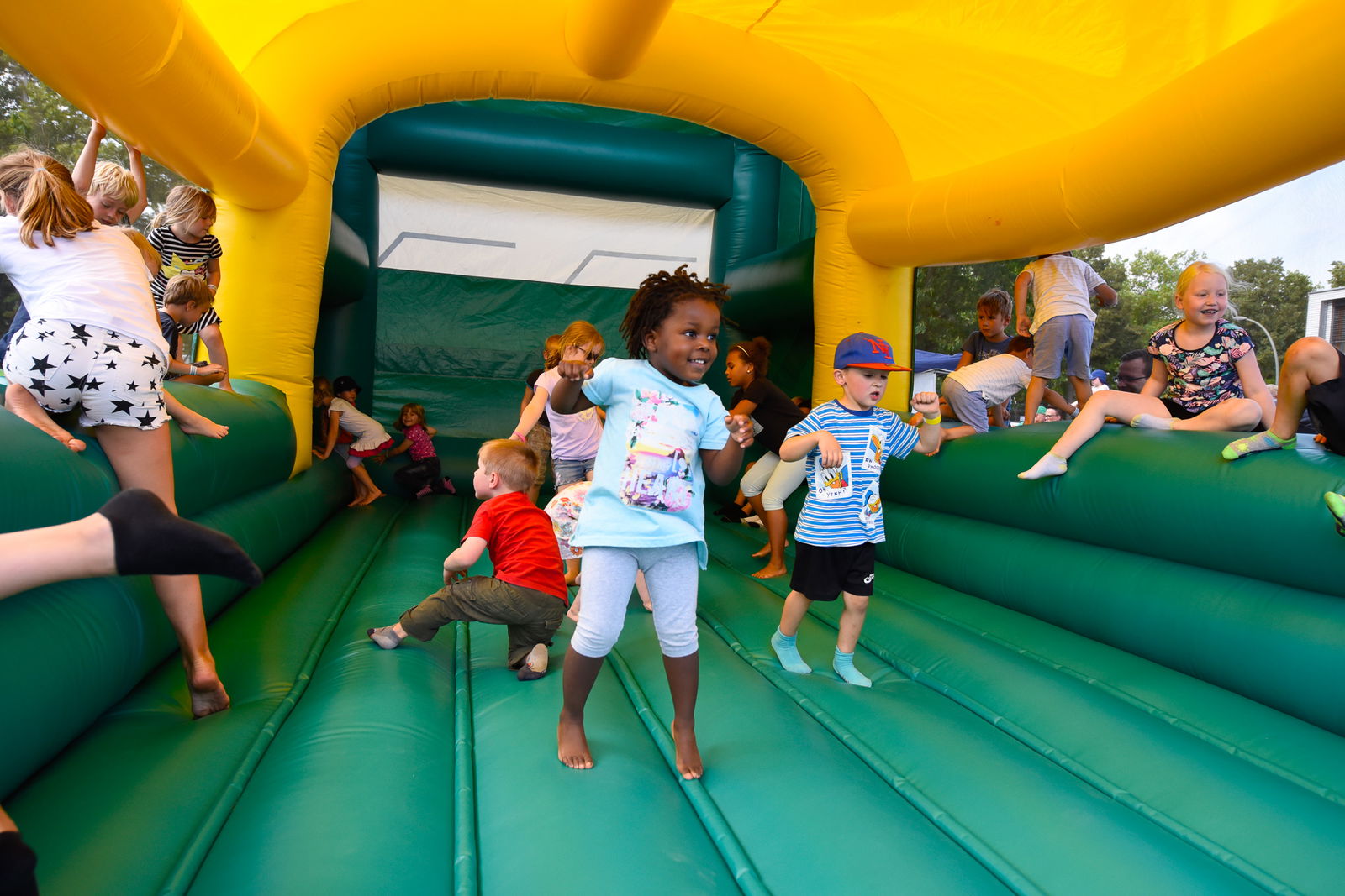 Das innere einer Hüpfburg. Mehrere kleine Kinder hüpfen auf einer mit Luft befüllten, grünen Fläche. Auf dem Rand der Hüpfburg sitzen, auf beiden Seiten, Kinder und gucken zu. Die Hüpfburg hat ein gelbes Dach.  