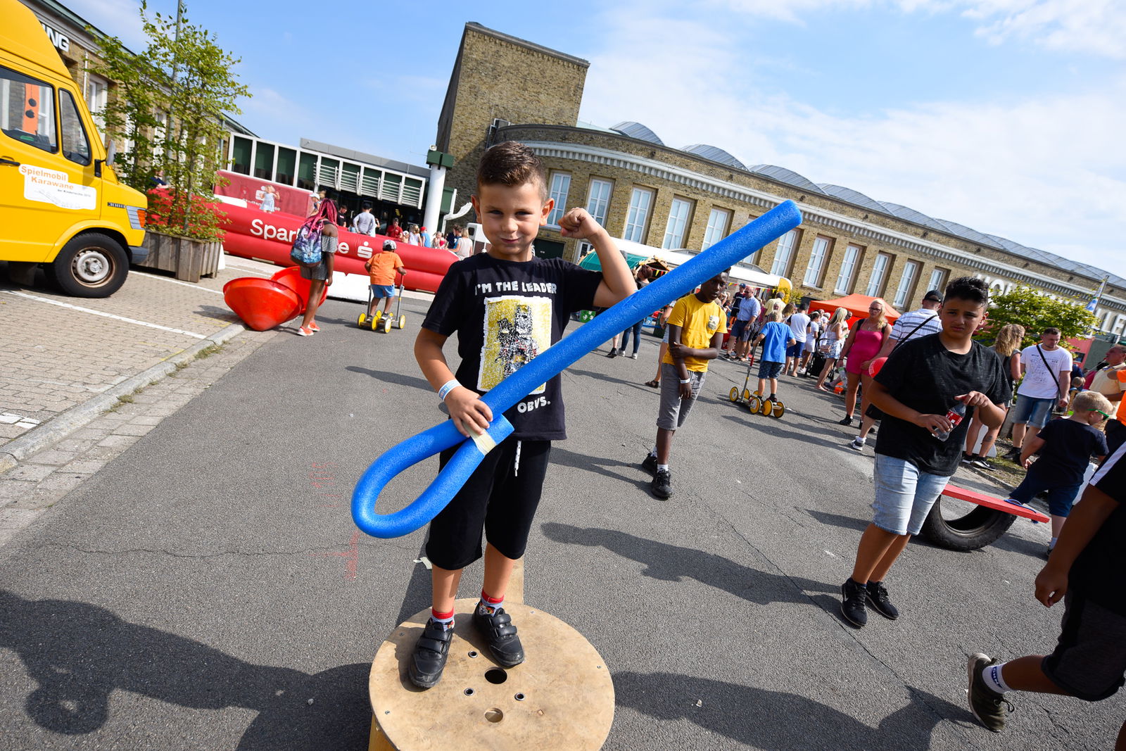 Ein kleiner Junge steht auf einer runden Holzplatte. In der Hand hält er einen blauen, langen Stab aus Styropor, der an einem Ende zu einer Öse geformt ist. Der Junge lächelt in die Kamera.
