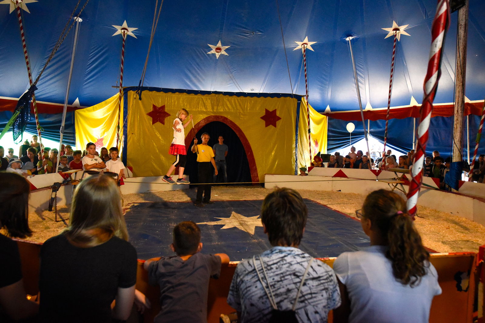 In einer Zirkusmanege balanziert ein junges Mädchen von links nach rechts über ein Seil. Ein Dompteur, der einen Stab in der Hand hält, begleitet sie. Auf den Rängen sitzen Zuschauer.