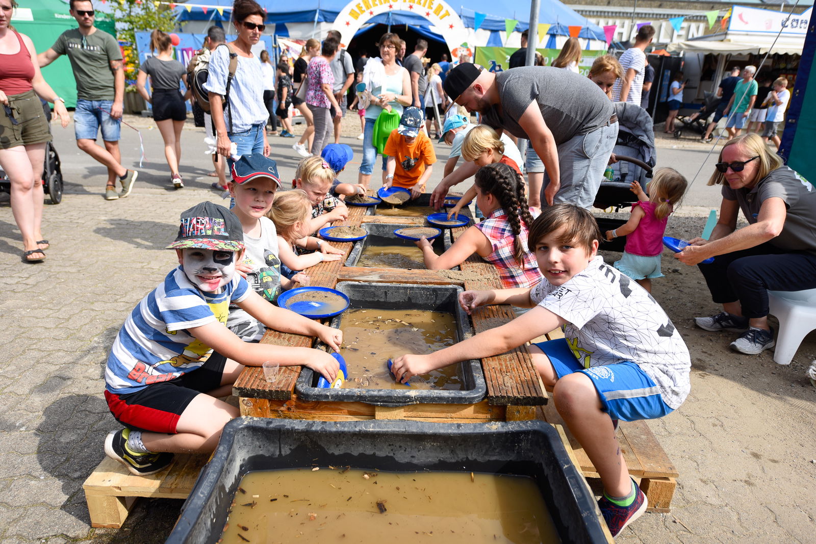 Mehrere Wasserbecken sind hintereinander angeordnet und mit Wasser gefüllt. Auf beiden Seiten der Bassins sitzen Kinder die mit Frisbees Sand vom Grund der Behälter sammeln und auf etwas untersuchen.