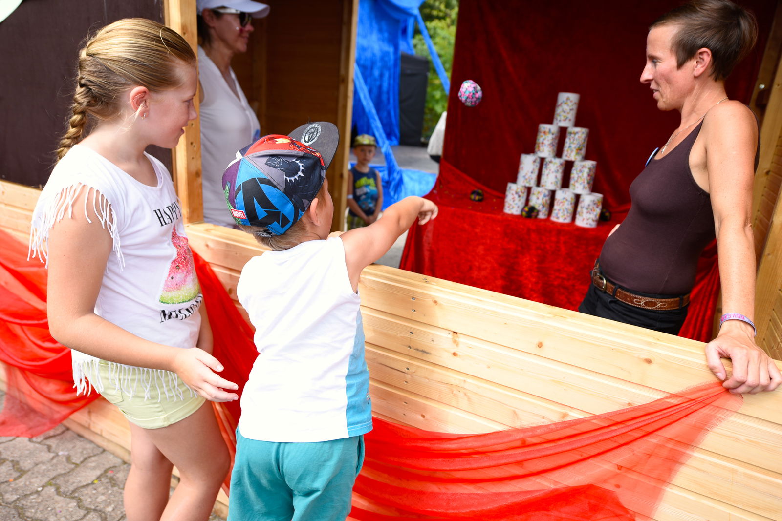 An einem Dosenwurfstand stehen nebeneinander ein junges Mädchen und ein kleiner Junge. Der Junge wirft gerade einen Ball auf einen Dosenstapel. Rechts daneben steht eine Verkäuferin und schaut zu. 