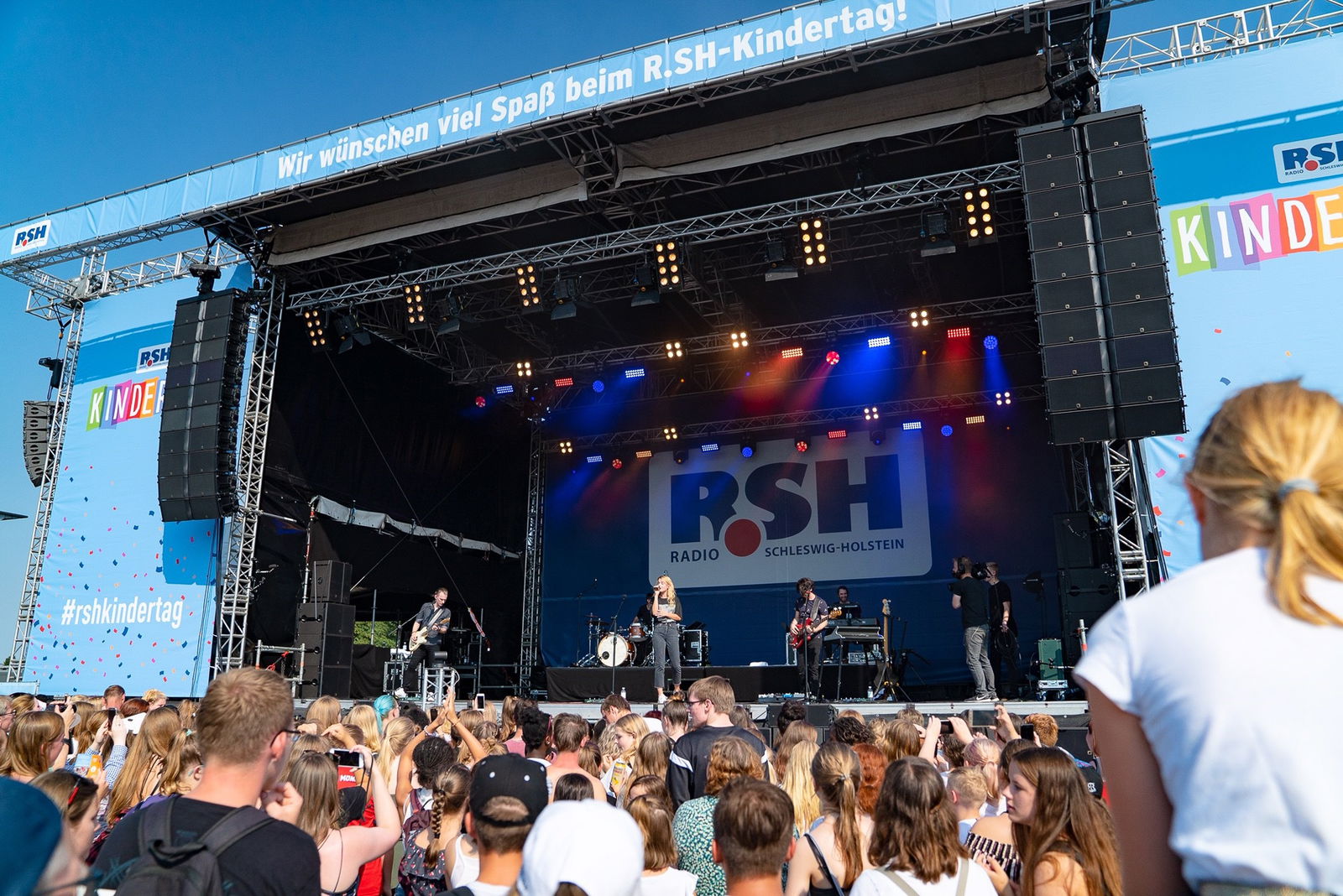 Eine Menschenmenge steht vor einer großen Bühne auf der gerade eine Band spielt und eine Frau in ein Mikrofon singt. 