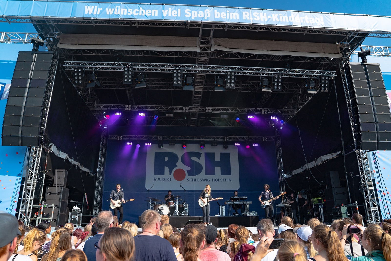 Viele Menschen stehen vor einer großen Bühne auf der gerade eine Band spielt.