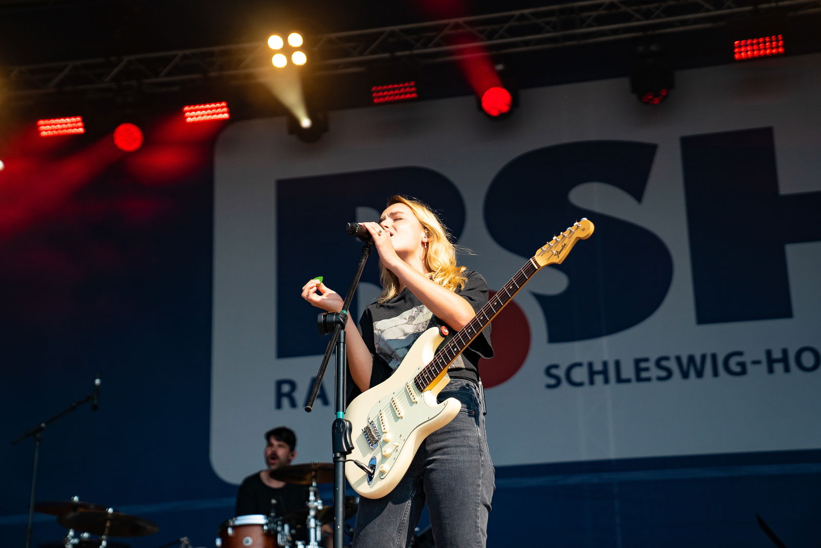 Eine Frau steht auf einer Bühne. Sie hat eine EGitarre vor sich umgebunden und singt in ein Mikrofon.