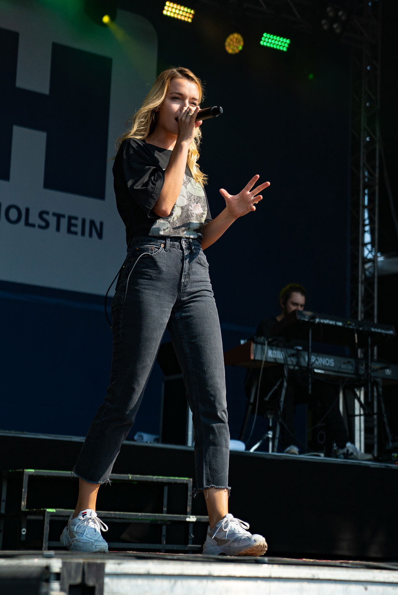 Eine Sängerin mit langen blonden Haaren steht auf einer Bühne und singt in ein Mikrofon.
