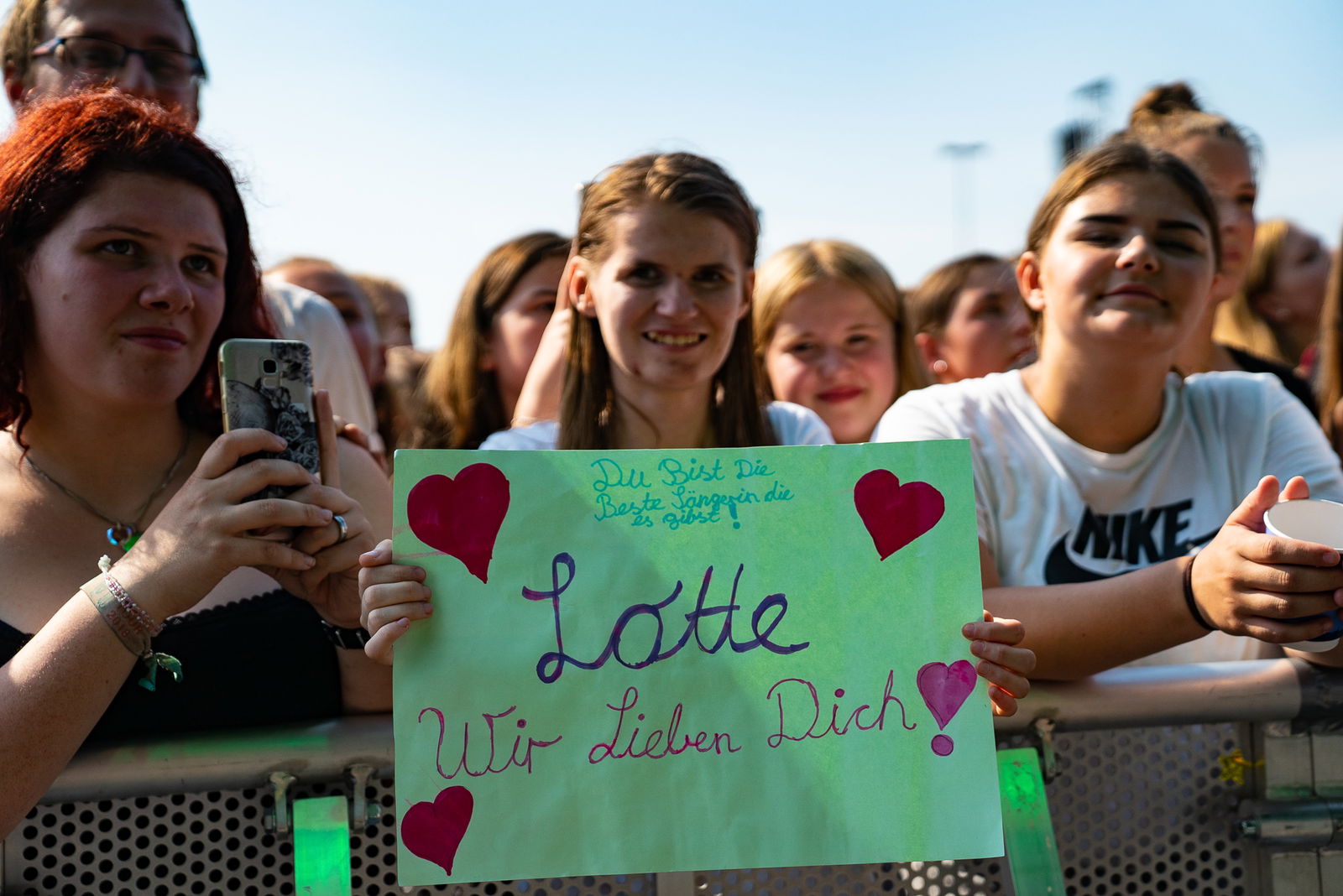 Drei Fans stehen an einem Absperrgitter einer Bühne, halten ein Schild mit der Aufschrift Lotte wir lieben dich und Herzen in der Hand.