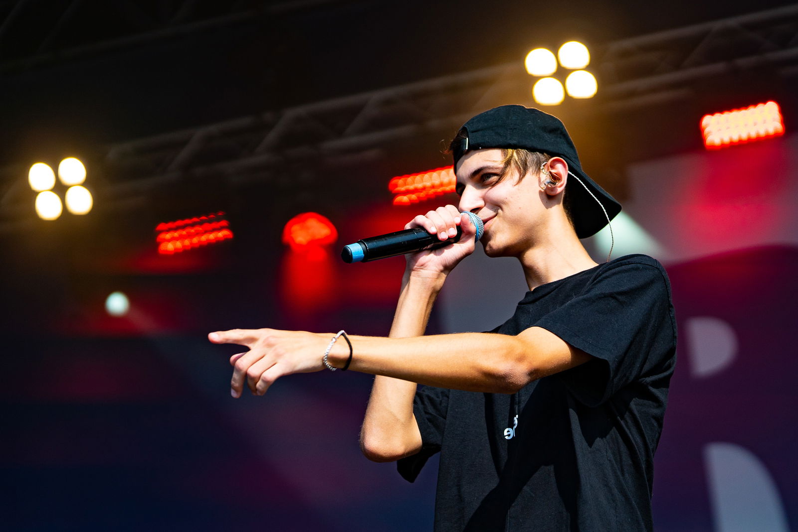 Ein junger Mann steht rechts im Bild, mit Blick nach links. Er singt in ein Mikrofon. Der linke Arm ist gestreckt, mit dem Zeigefinger zeigt er nach vorne.