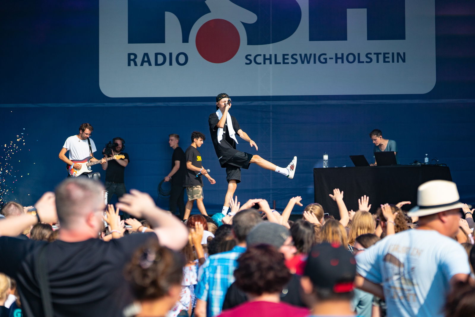 Eine Menschenmenge steht vor einer Bühne auf der eine Band steht. Ein junger Mann singt in ein Mikrofon und hebt dabei sein rechtes Bein seitlich nach rechts in die Höhe. 