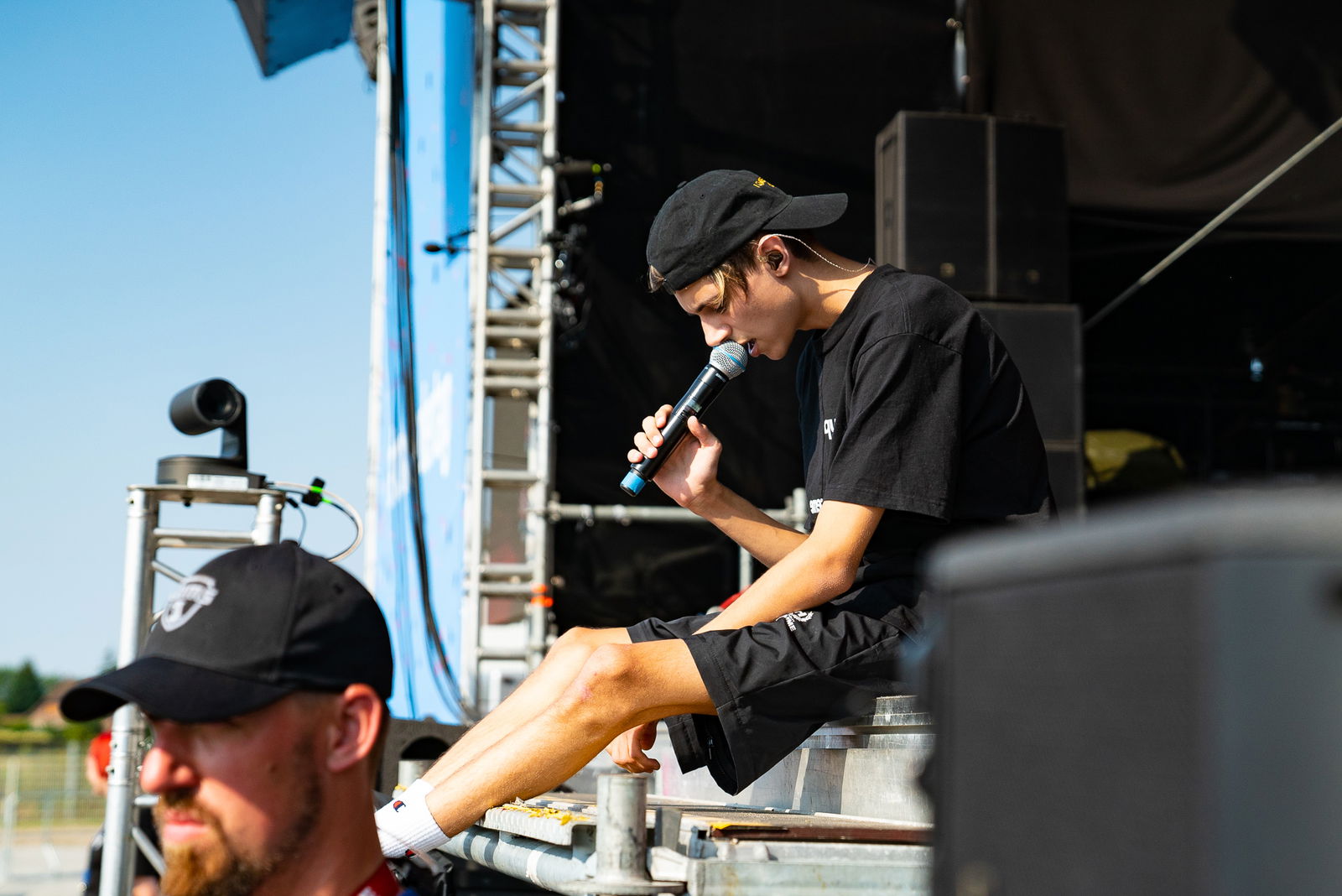 Ein junger Mann sitzt auf dem Rand einer Bühne und singt in ein Mikrofon. links vor ihm ist der Kopf von einem Mann des Sicherheitspersonals zu sehen der nach links blickt.