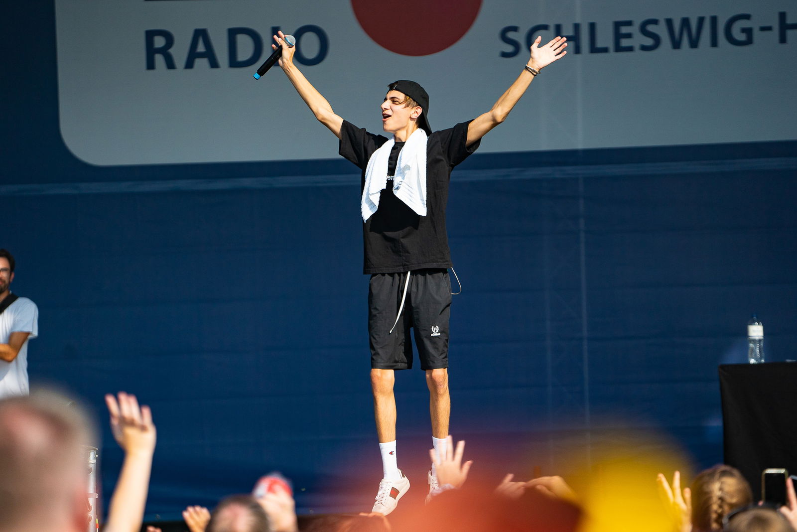 Ein junger Mann springt auf einer Bühne in die Luft und hebt dabei seine Arme in die Höhe. In der rechten Hand hat er ein Mikrofon. Vor der Bühne stehen menschen die ihm zujubeln.