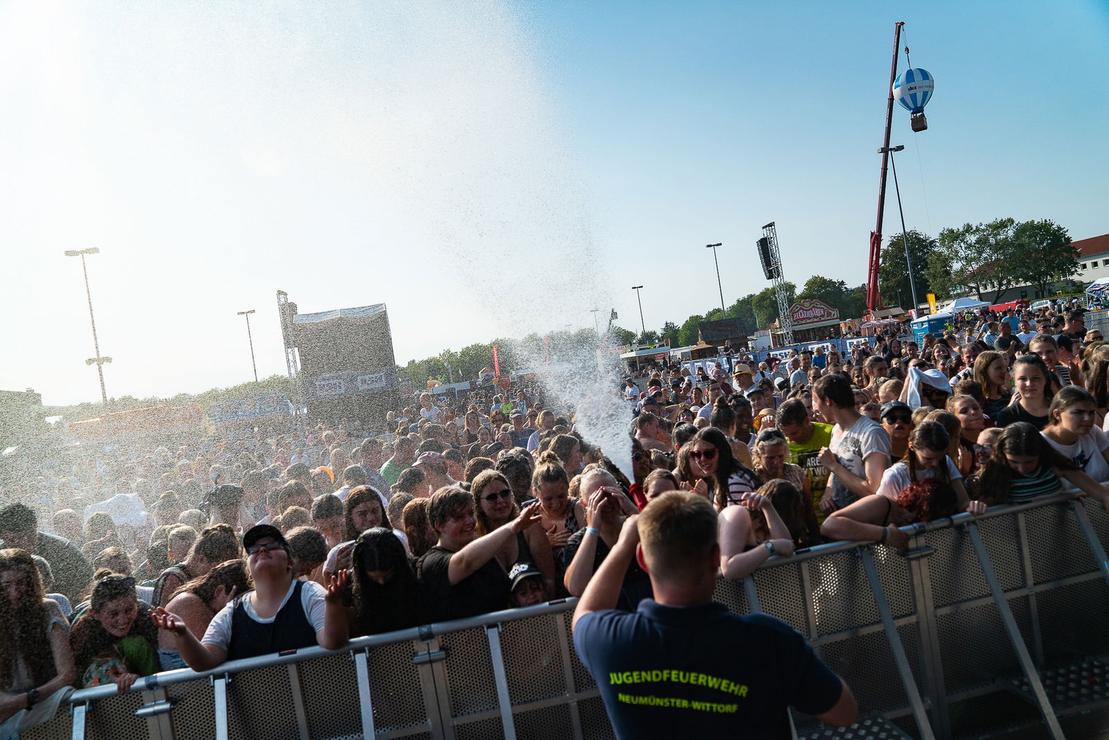 Ein Feuerwehrmann steht mit dem Rücken zur Kamera. Er hat einen Feuerwehrschlauch in der Hand aus dem Wasser spritzt, mit dem er die vor sich stehende Menschenmenge nass spritzt. Zwischen ihm und der Menschenmenge ist ein Absperrgitter aufgebaut.