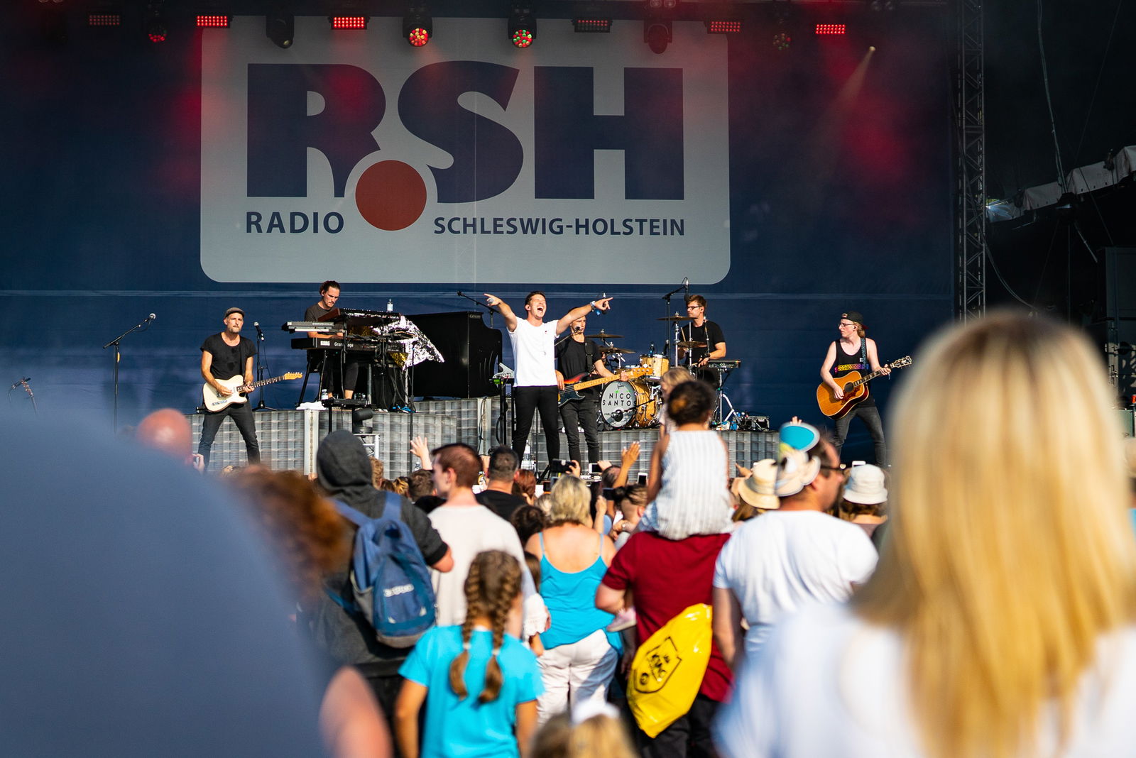 Ein Mann steht zusammen mit einer Band auf einer großen Bühne und streckt die Arme nach oben. Vor der Bühne steht eine Menschenmenge die ihm dabei zusieht.
