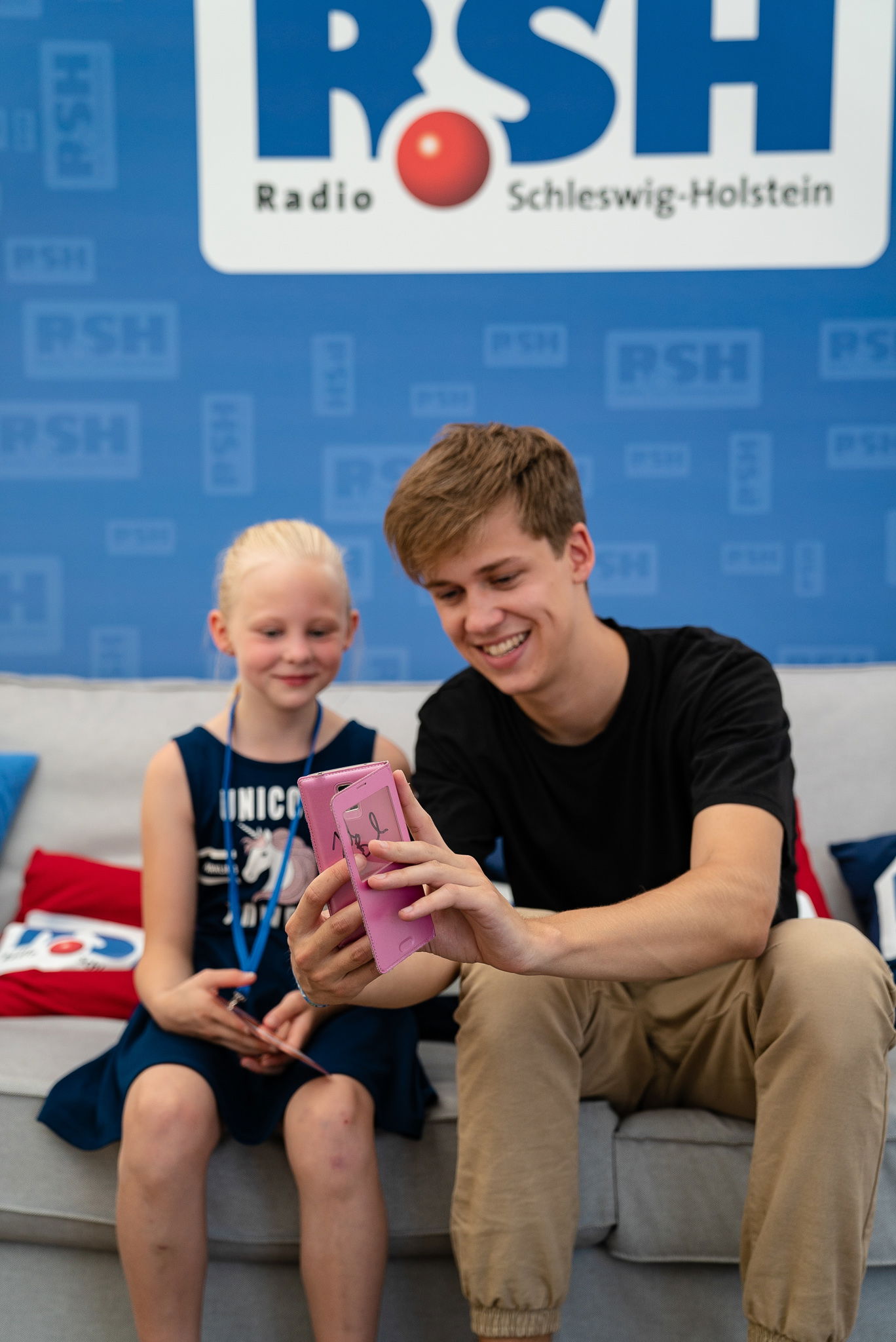Ein junger Mann und ein kleines Mädchen sitzen gemeinsam auf einem Sofa. Der Mann hält ein rosanes Handy in der Hand auf das beide schauen.