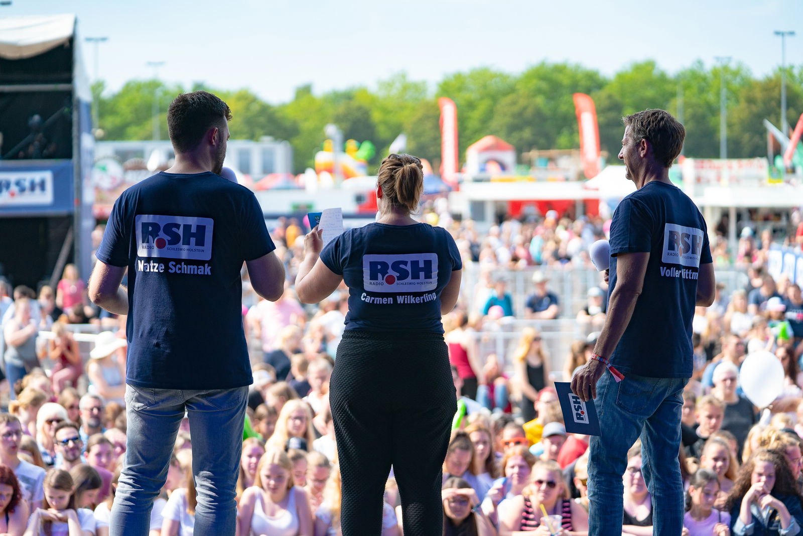 Eine Frau steht gemeinsam mit zwei Männern, die links und rechts neben ihr stehen, auf einer großen Bühne. Die Personen stehen mit dem Rücken zur Kamera auf und blicken auf eine Menschenmenge die vor der Bühne steht.