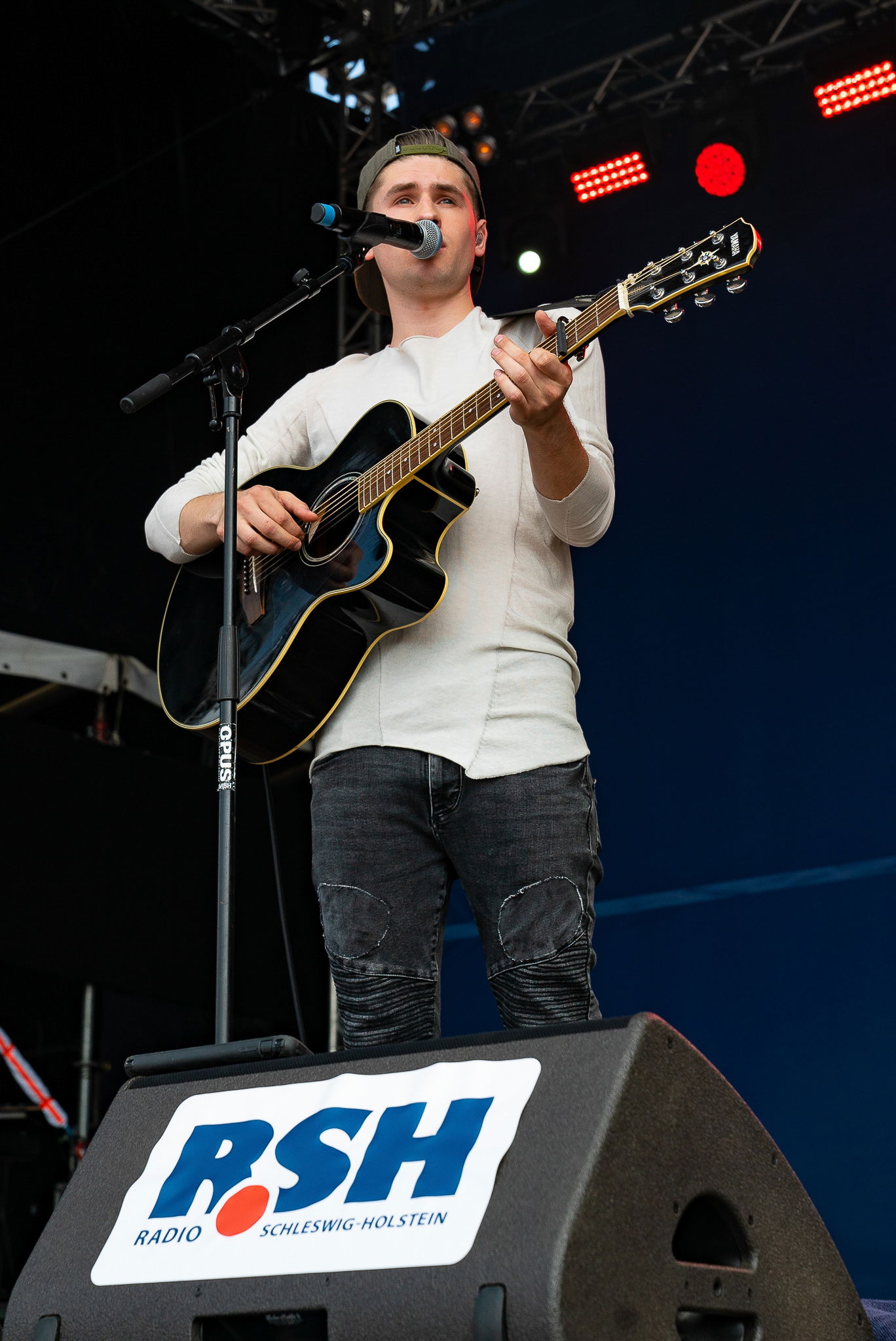 Ein junger Mann steht auf einer Bühne und spielt auf einer Gitarre während er in ein Mikrofon singt, was vor ihm an einem Mikrofonständer angebracht ist.