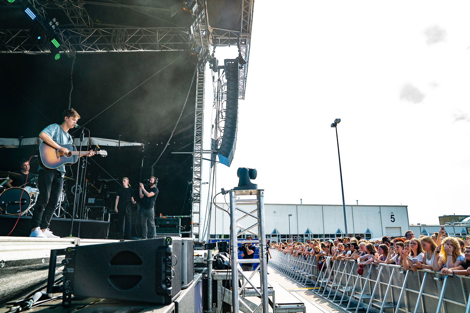 Ein junger Mann steht auf der Bühne. Er spielt auf einer Gitarre, singt in ein Mikrofon und schaut in eine Menschenmenge, die rechts gegenüber der Bühne steht.