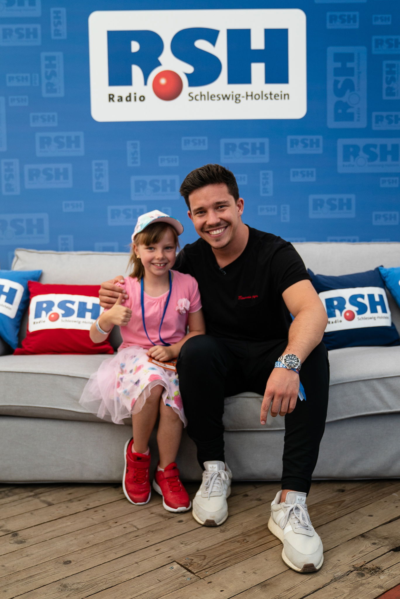 Der Sänger Nico Santos sitzt rechts neben mit einem jungen Mädchen auf einer Couch. Gemeinsam schauen sie in die Kamera.