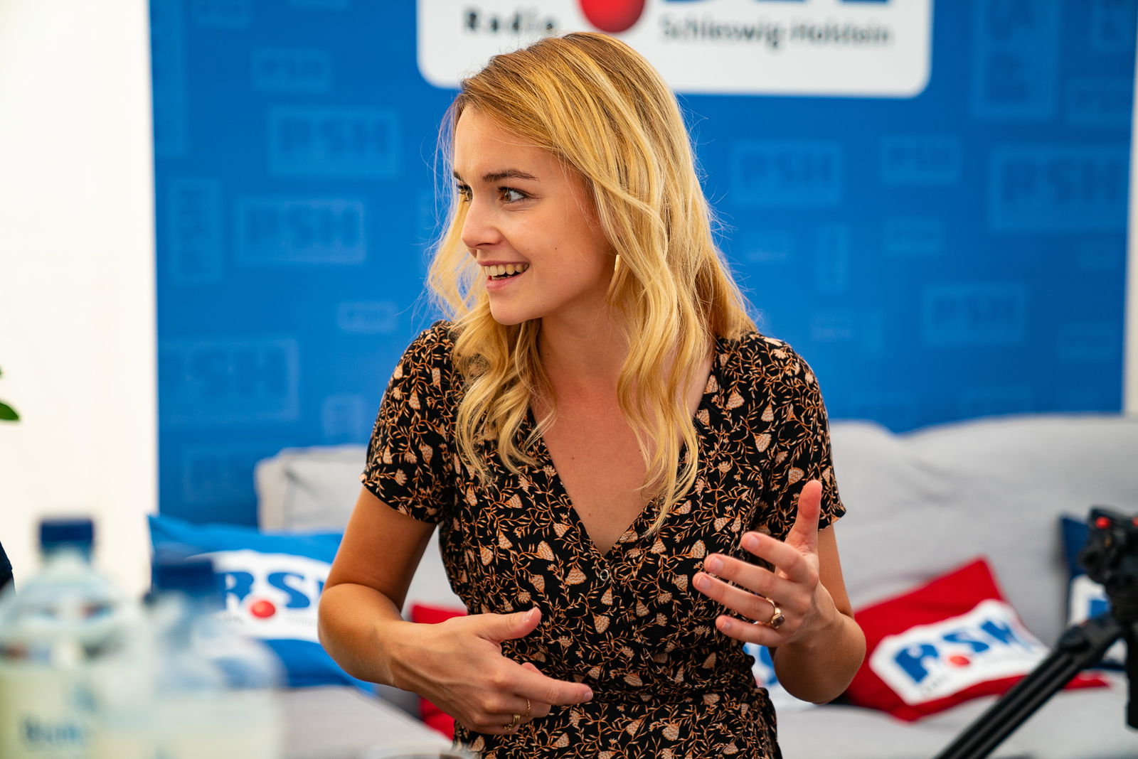 Eine junge Frau mit langen blonden Haaren sitzt der Kamera zugewandt an einem Tisch. Sie schaut nach links und gestikuliert mit den Händen. 