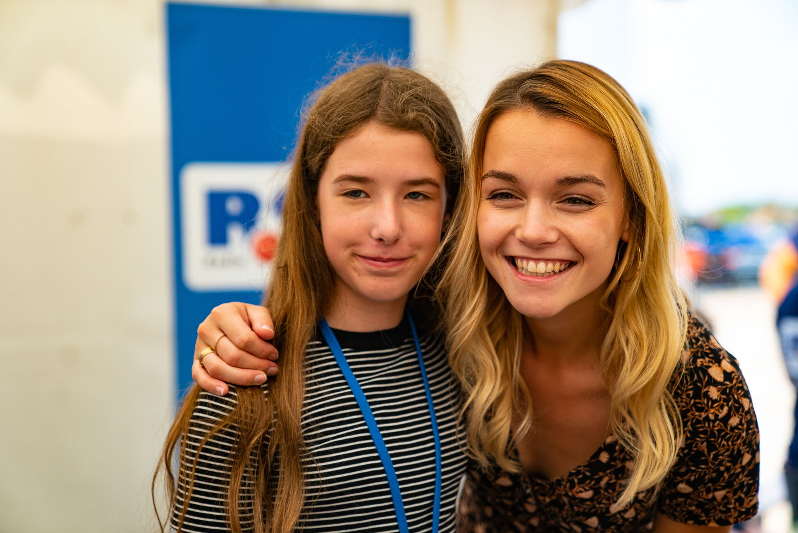 Eine junge Frau mit langen blonden Haaren hat den Arm um die Schulter eines jungen Mädchens gelegt, das rechts neben ihr steht. Beide lächeln in die Kamera.