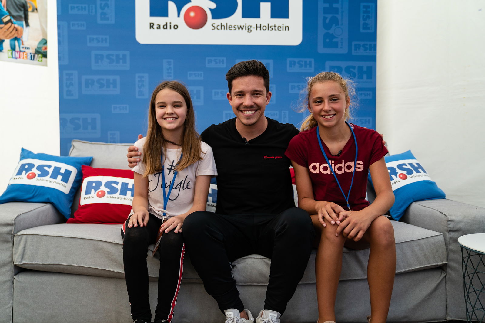 Der Sänger Nico Santos sitzt gemeinsam mit zwei jungen Mädchen auf einer Couch. Die Mädchen sitzen links und rechts von ihm.