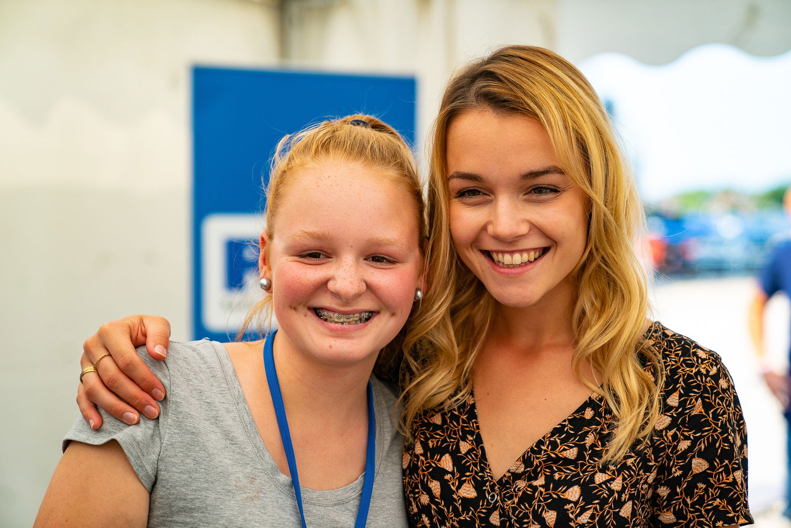 Eine junge Frau mit langen blonden Haaren hat den Arm um die Schulter eines Mädchens gelegt, das rechts neben ihr steht. Beide lächeln in die Kamera.