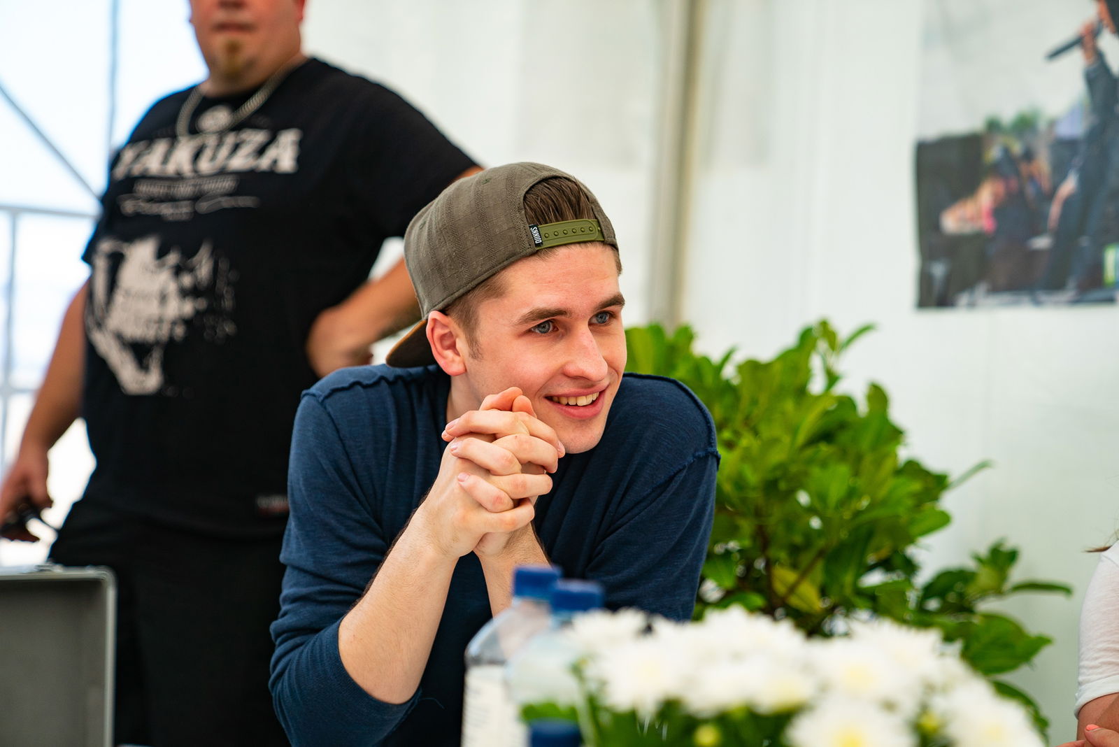 Ein junger Mann, der eine Kappe mit dem Schirm nach hinten trägt, sitzt der Kamera zugewandt an einem Tisch. Er sieht nach rechts, hat die Arme abgestützt und die Hände gefaltet. 