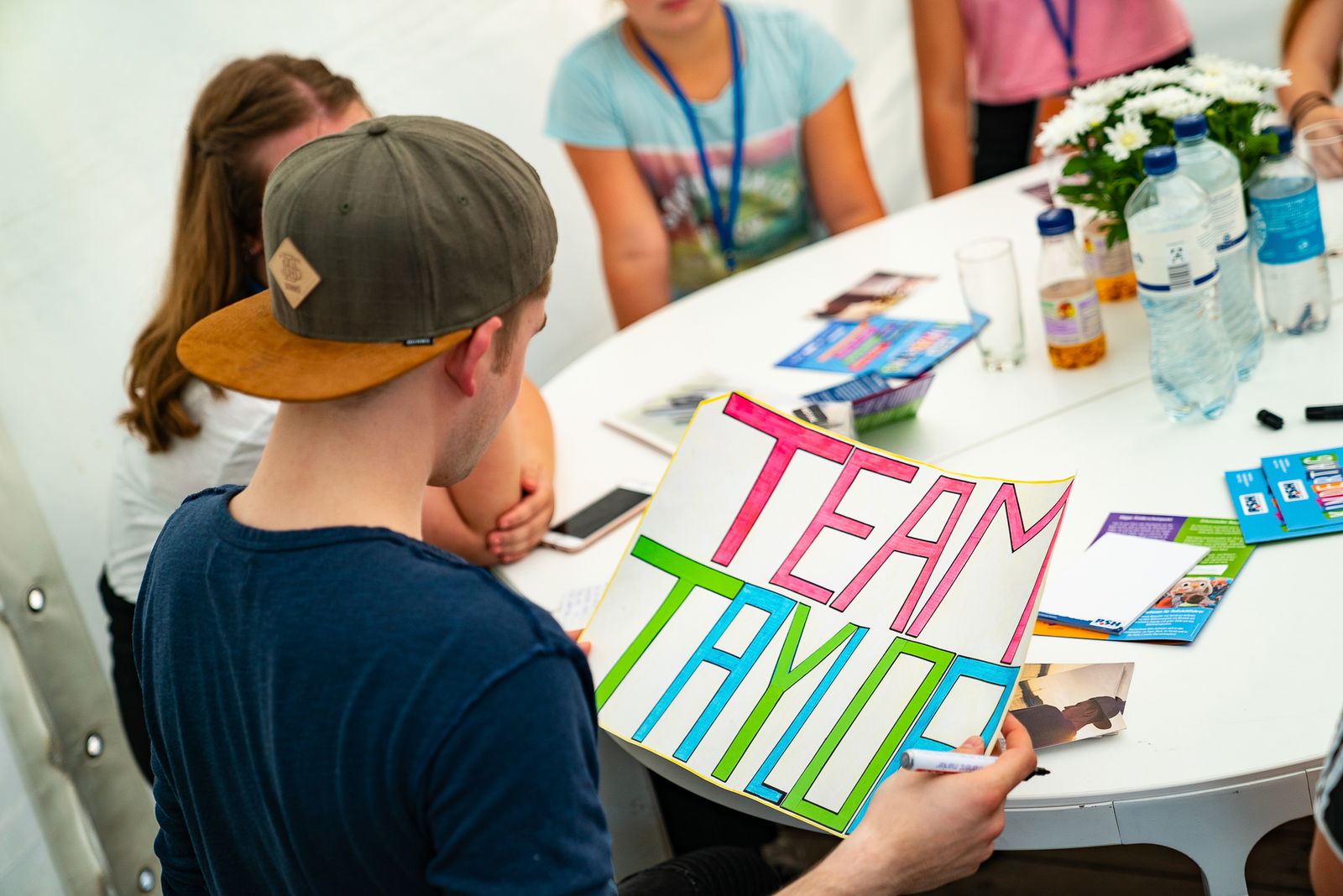 Ein junger Mann, der an einem Tisch sitzt, hält ein Plakat in der Hand auf dem Team Taylor steht. Er sitzt mit dem Rücken zur Kamera, links im Bild. 