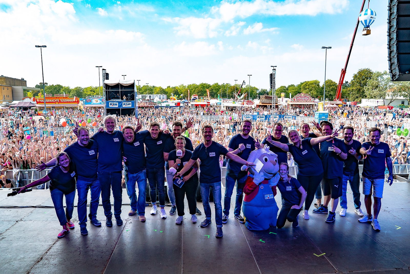 Eine Gruppe von Menschen steht auf einer großen Bühne und jubelt in die Kamera. Im Hintergrund ist eine Menschenmenge zu sehen die auf die Bühne sieht.