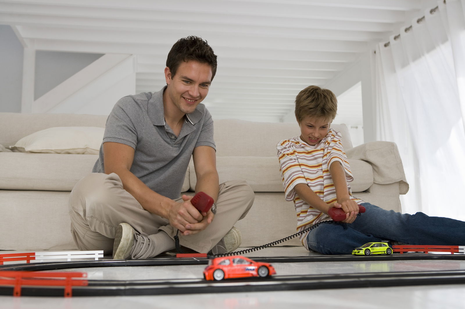 Vater und Sohn sitzen vor einer Couch, haben Controller in der Hand und spielen mit einer Carrerabahn