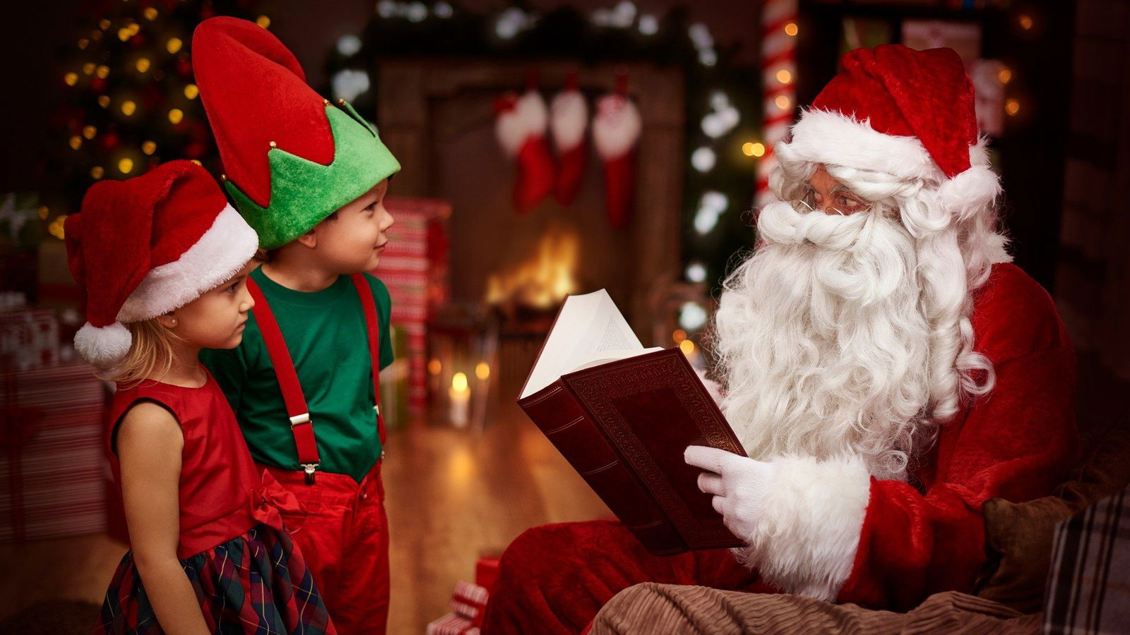 Ein kleines Mädchen und ein kleiner Junge stehen vor einem Mann der auf einem Stuhl sitzt, ein Weihnachtskostüm und einen langen weißen Bart sowie eine rot weiße Mütze auf dem Kopf trägt. In seinen Händen hält er ein aufgeklapptes großes Buch.