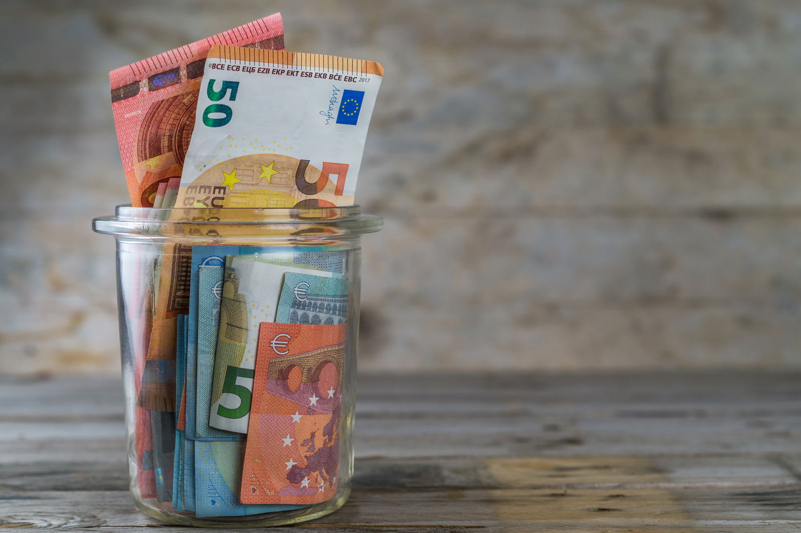 In einem Glas stecken viele Euroscheine, das auf einer Holzplatte steht.