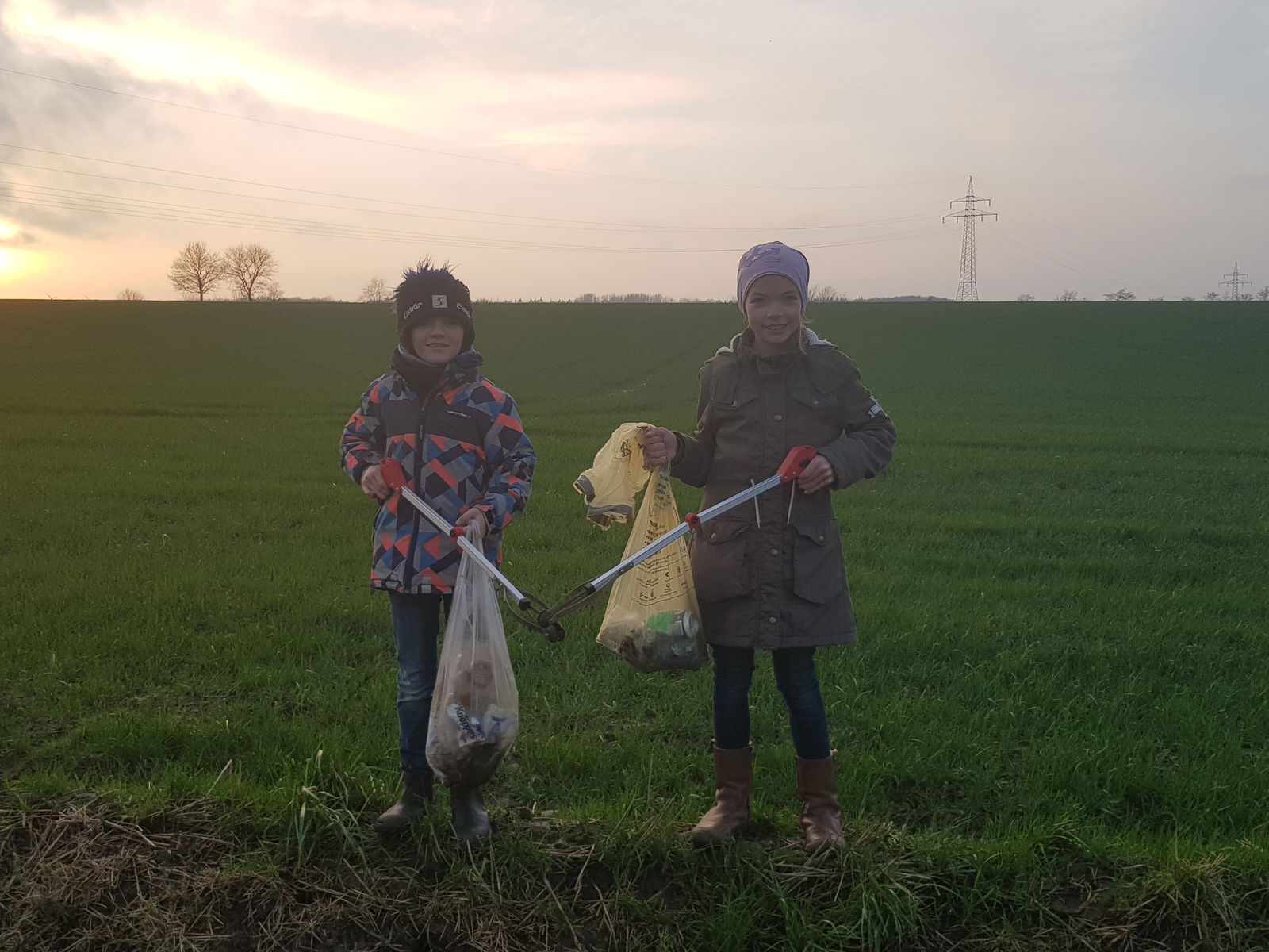 Ein Junge und ein Mädchen stehen auf einer Wiese. Sie sind dick angezogen. In ihren Händen halten sie je einen Müllsack und je eine Zange an einem Stab zum Müll aufsammeln. 
