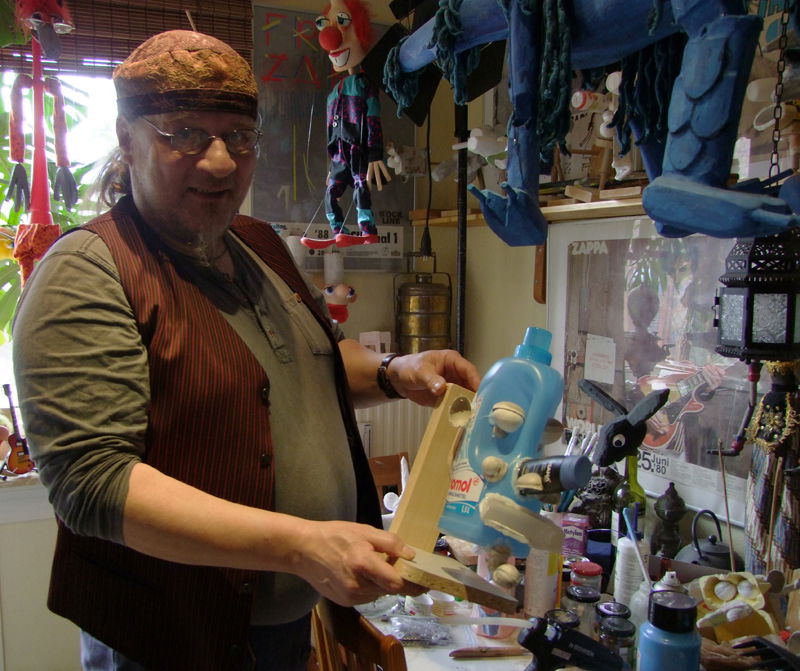 Ein Mann steht vor einem Tisch, auf dem viele Materialien liegen. In seinen Händen hält er zusammengebastelte Utensilien aus Müll. 