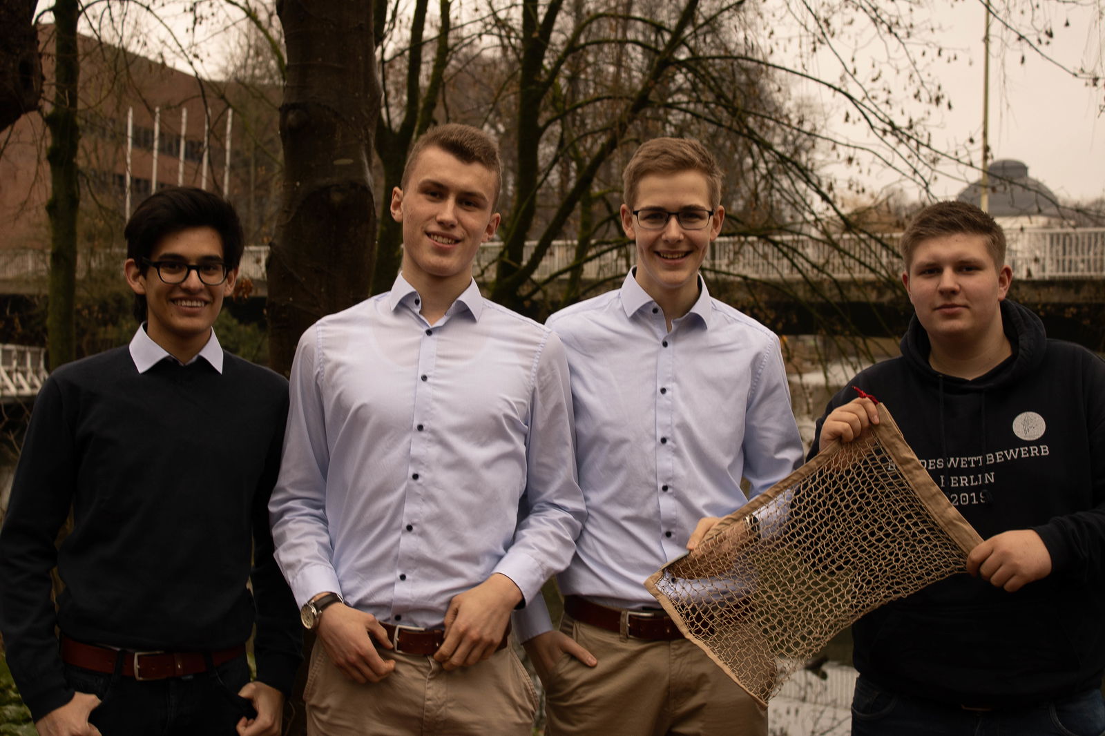 Vier Jungs stehen nebeneinander vor Bäumen und gucken in die Kamera. Die beiden, die rechts stehen, halten vor sich ein Netz in den Händen.