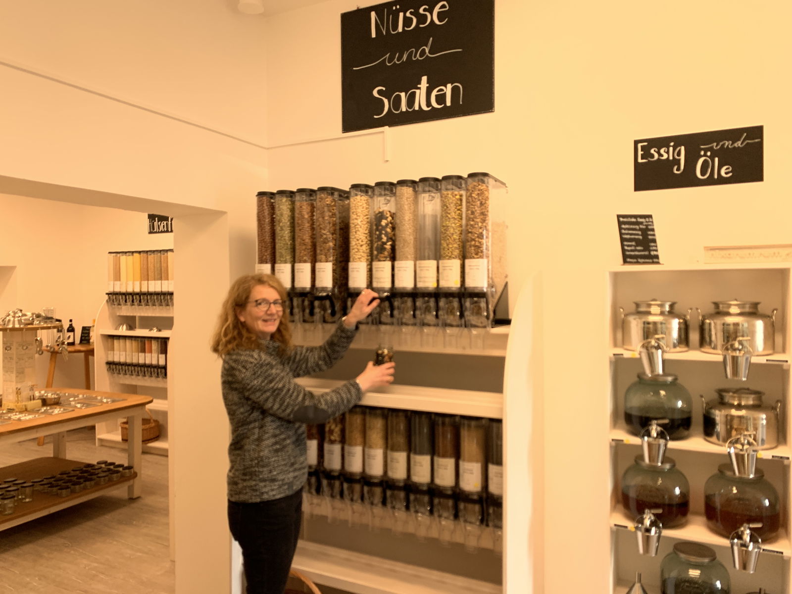 Eine Frau steht vor einer Wand, an der viele Behälter hängen. In ihrer rechten Hand hält sie ein Gefäß und befüllt es mit Lebensmitteln.