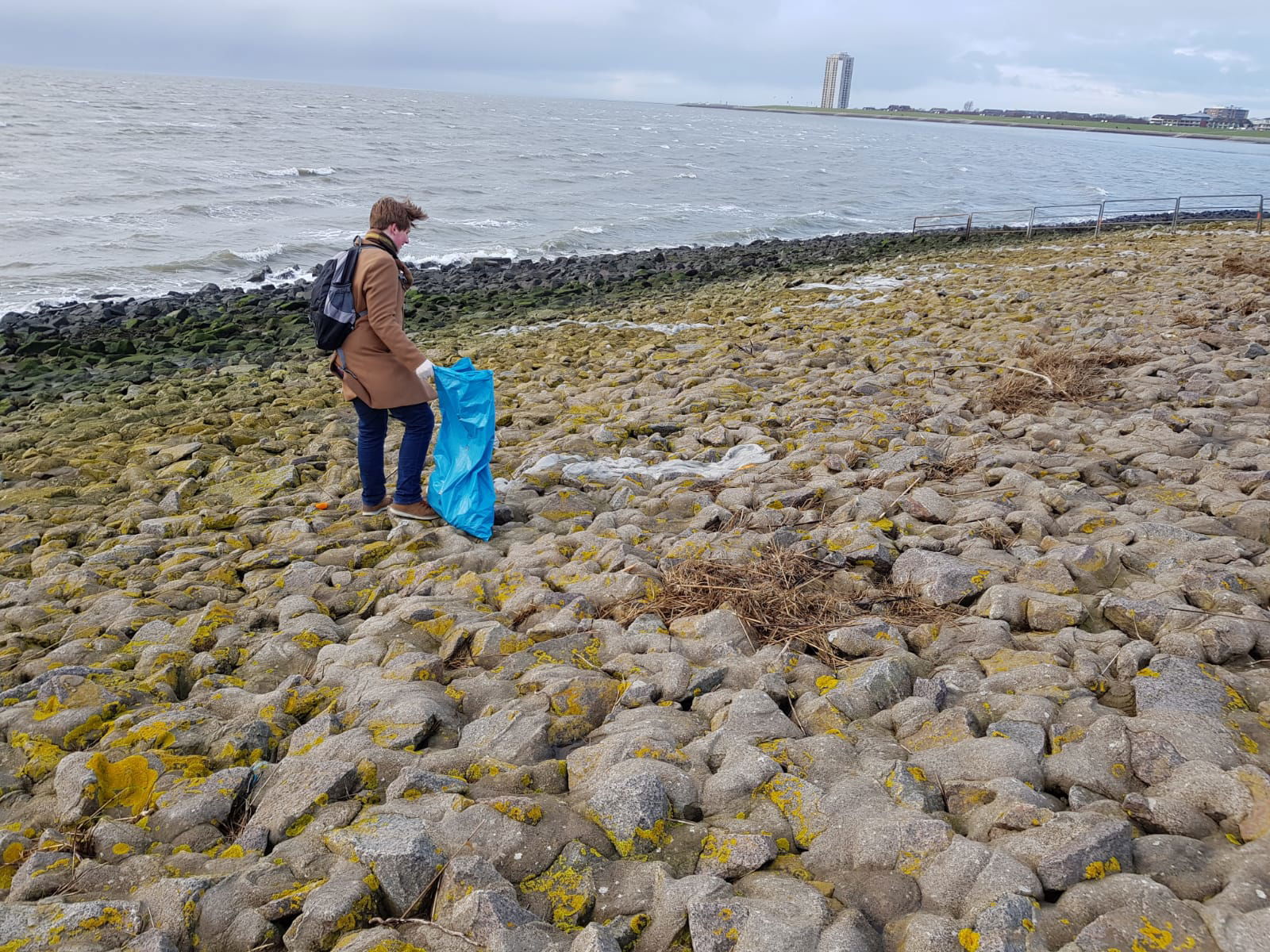 Ein Mann steht auf Steinen vor einem Meer und hat eine Mülltüte in seiner rechten Hand. 