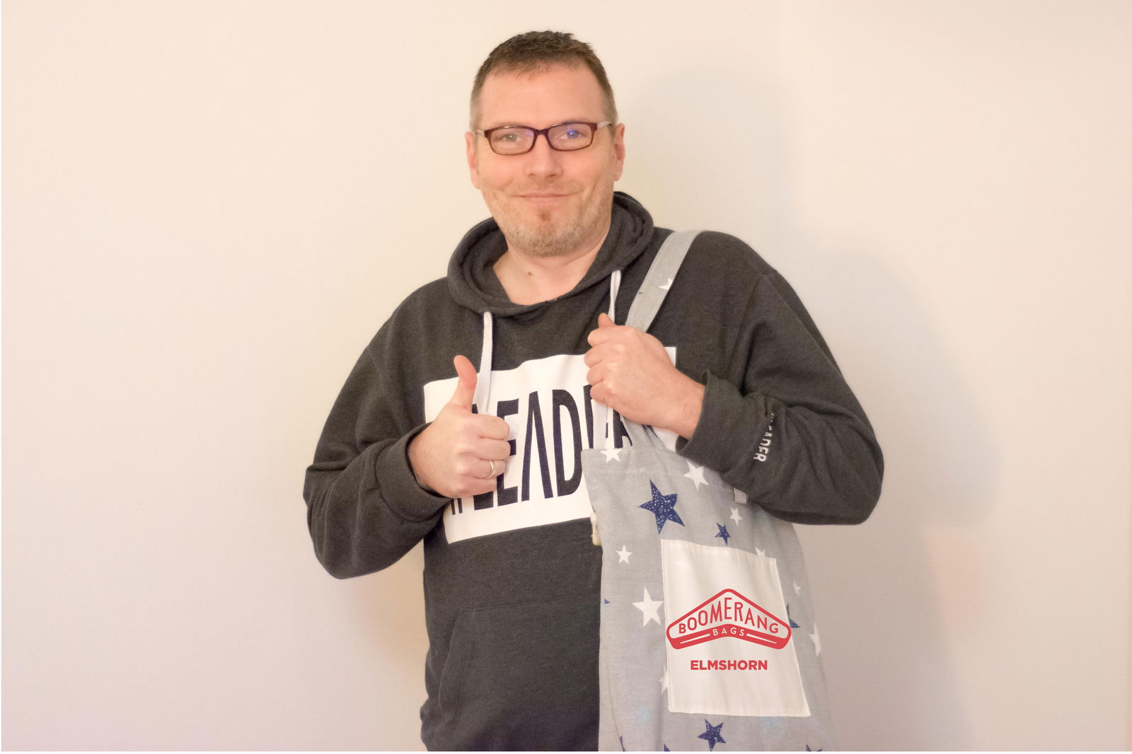 Ein Mann steht vor einer hellen Wand und hält seinen Daumen nach oben. In der linken Hand hat er eine Stofftasche mit der Aufschrift Boomerang Bags.